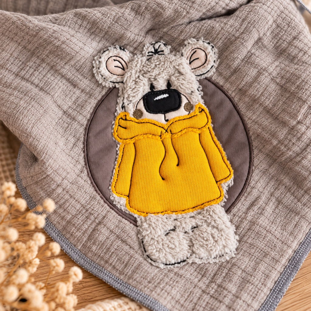 Nahaufnahme der Teddy-Stickerei im senfgelben Hoodie auf graubeigem Musselin